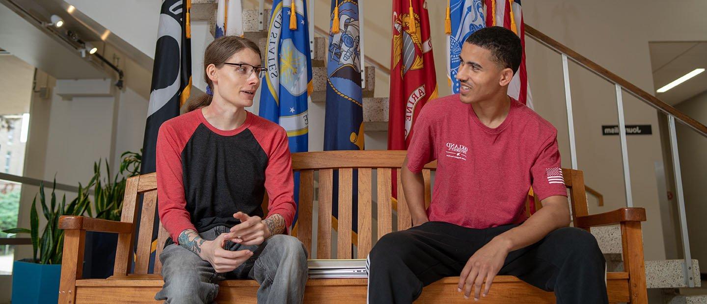 Two young men sit on a bench talking. There are a flags behind them.